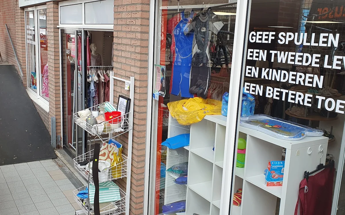 Winkelgevel met etalage vol tweedehandsartikelen, stellingen met kleding en speelgoed zichtbaar achter ramen
