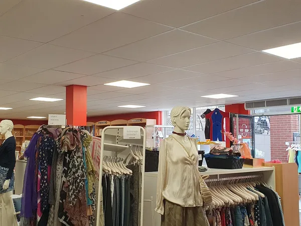 Rekken met tweedehands kleding, een etalagepop in beige blouse en bruine broek, en rode kledingdisplays in een kringloopwinke