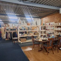 Winkelvloer met diverse afdeling: kledingrekken, houten tafels met stoelen, en volle stellingen met huishoudelijke artikelen 