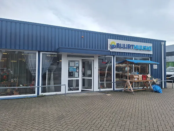 Blauwe gevel van kringloopwinkel Buurthuuske met winkelnaam, ingang en etalageruiten, voorkant met parkeerplaats