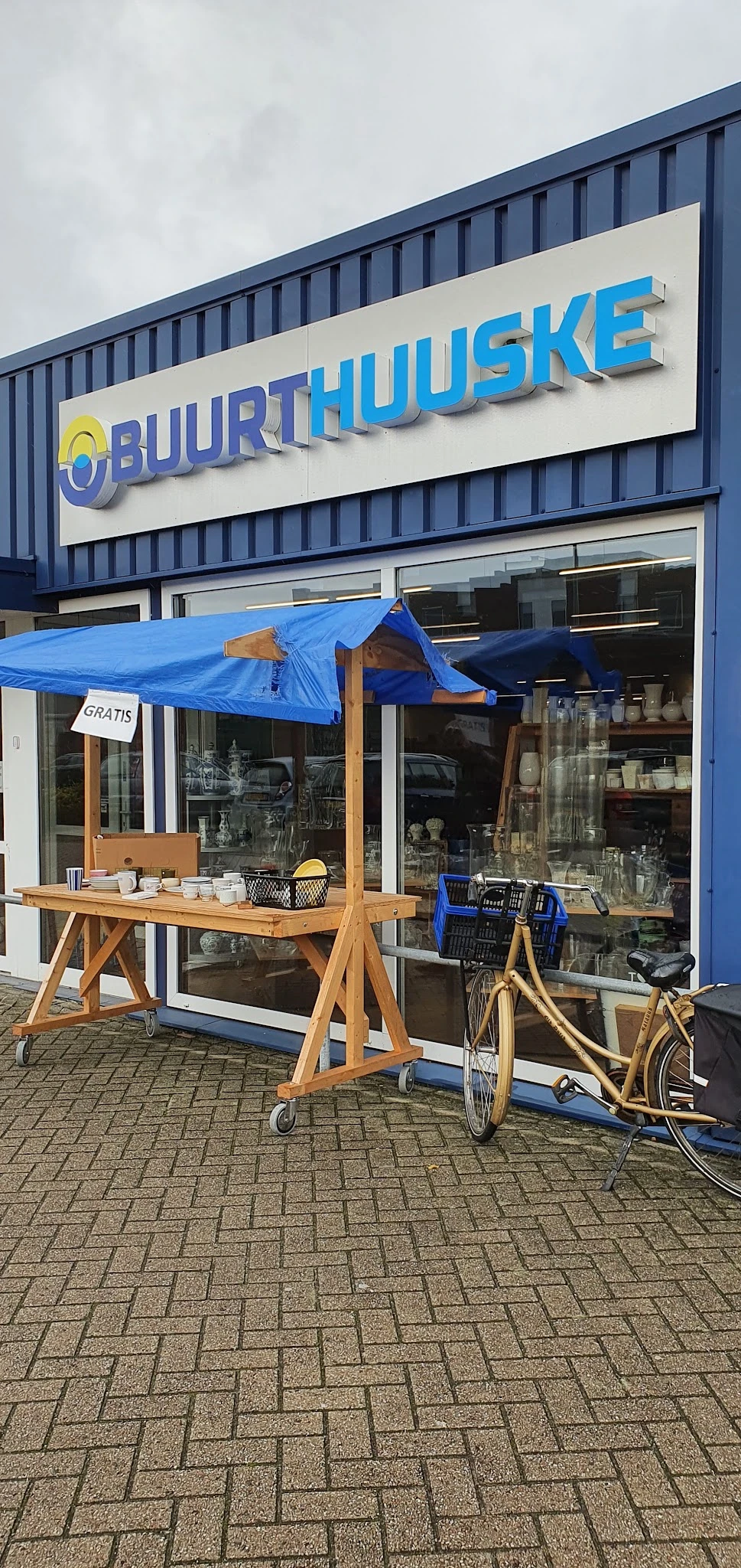 Buiten hoek van kringloopwinkel met blauwe luifel, houten toonbank met kopjes en een bord 'GRATIS'