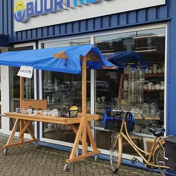 Buiten hoek van kringloopwinkel met blauwe luifel, houten toonbank met kopjes en een bord 'GRATIS'