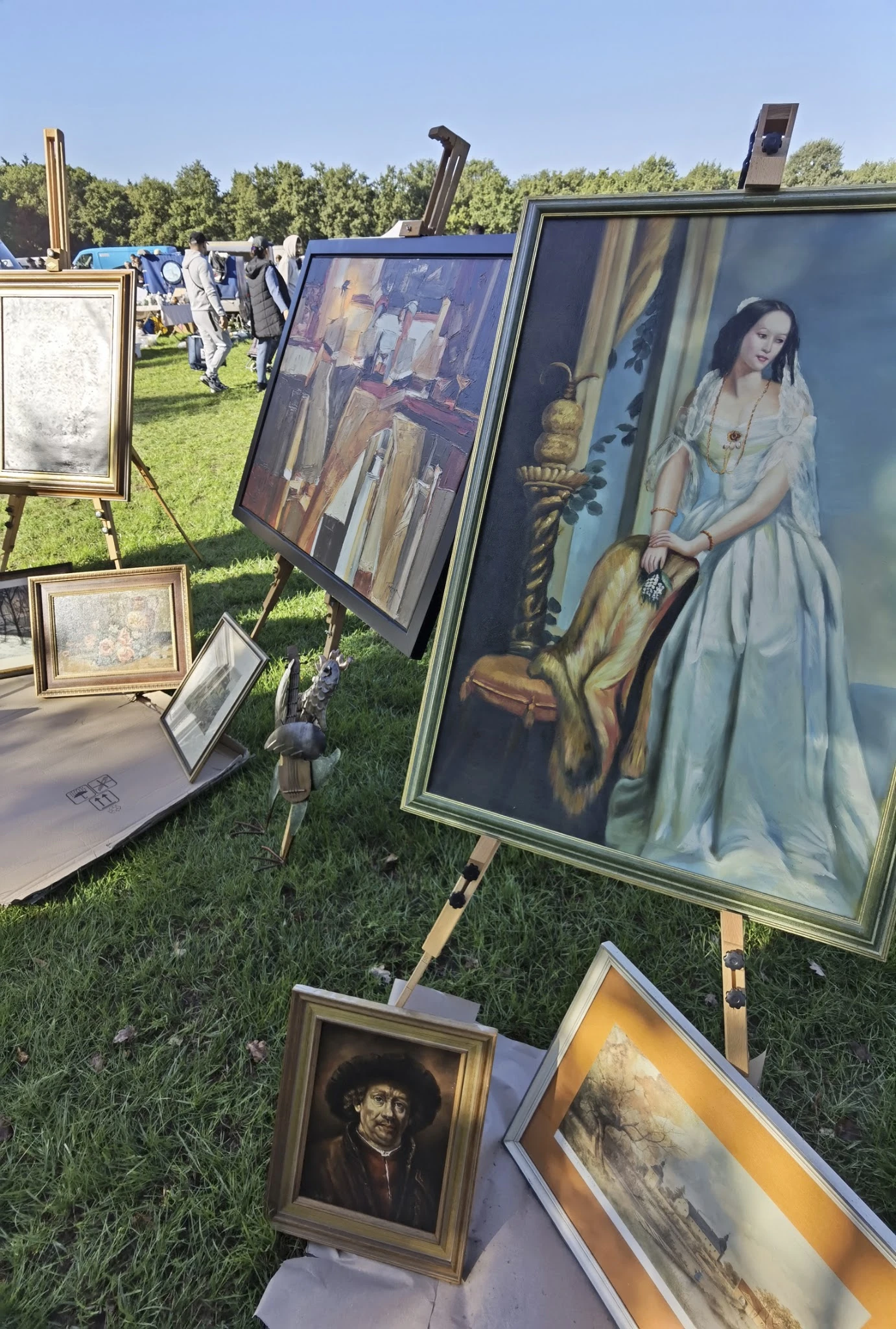Tweedehands kunstwerken en schilderijen op gras bij een kringloopmarkt, waaronder portret van vrouw en klassieke kunstwerken 