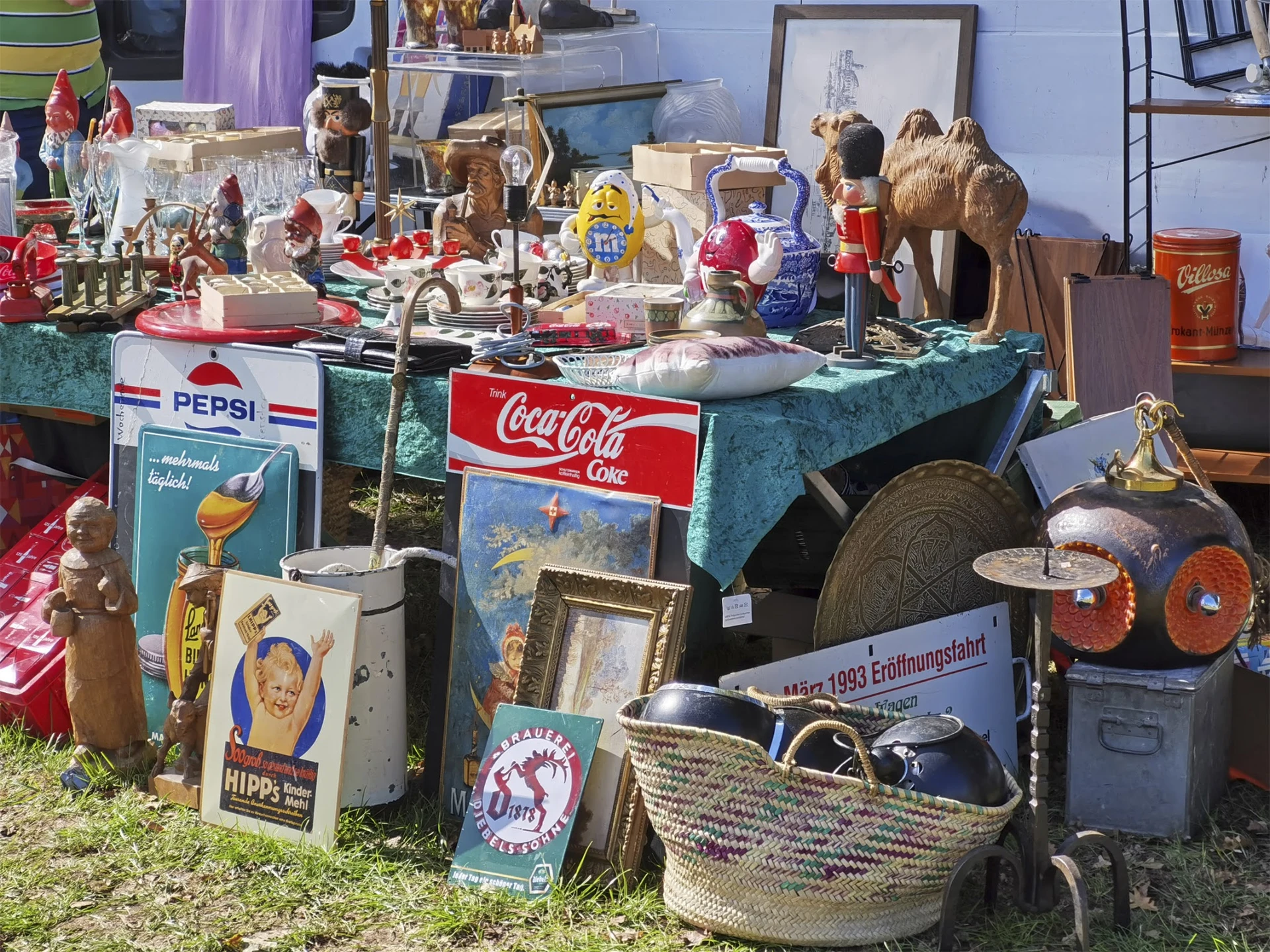 Carbootsale display met vintage items: Pepsi- en Coca-Cola borden, poppen, servies, sieraden en decoratie op blauw kleed