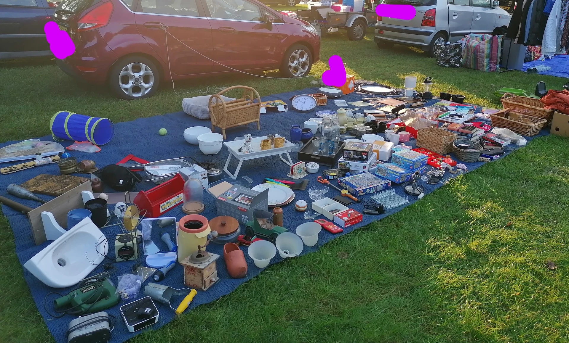 Uitgestalde tweedehands spullen op blauwe dekens op het gras tijdens carbootsale, met auto's en tenten op de achtergrond