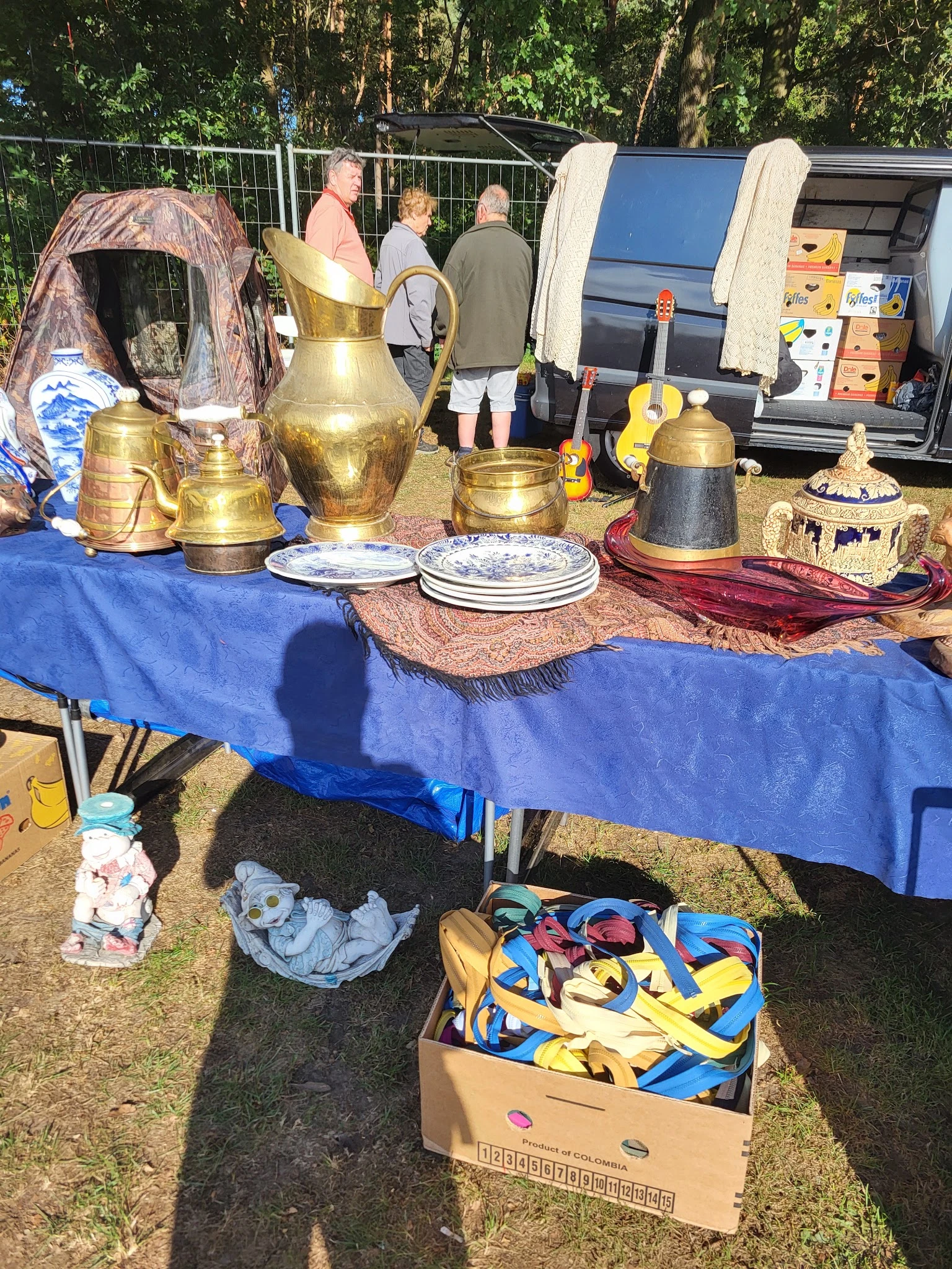 Uitgestalde tweedehands items op een blauwe tafel, waaronder koperen kannen, borden en decoratieve objecten