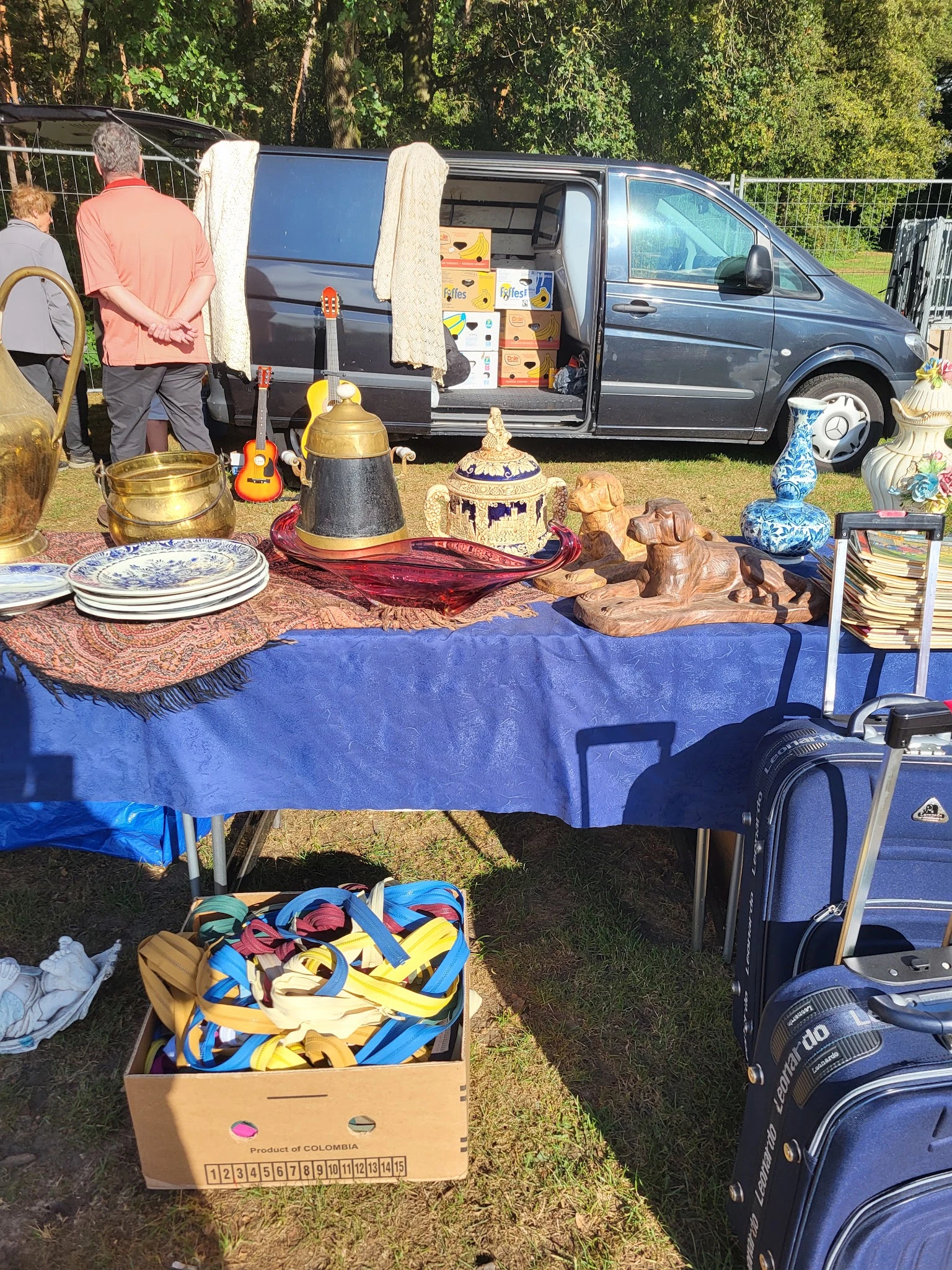 Uitgestalde tweedehands huishoudartikelen op een blauwe tafel bij een kringloopwinkel carbootsale, waaronder servies, kandela