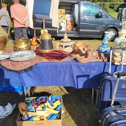 Uitgestalde tweedehands huishoudartikelen op een blauwe tafel bij een kringloopwinkel carbootsale, waaronder servies, kandela