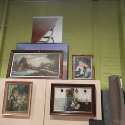 Uitgestalde schilderijen en kunstwerken in verschillende formaten en framesoorten tegen gele wand