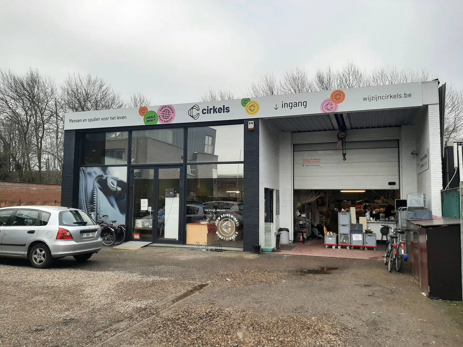 Voorkant van kringloopwinkel Cirkels met witte gevel, grote ramen en ingang, parkeerplaats met auto ervoor