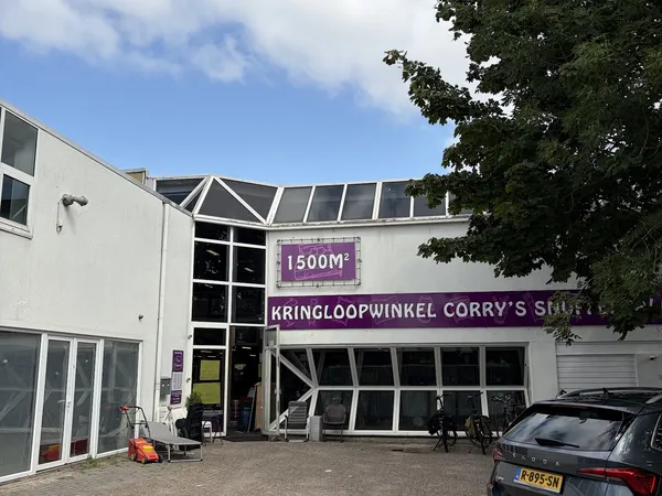 Witte gevel van kringloopwinkel met paarse borden en glazen pui, enkele auto geparkeerd ervoor