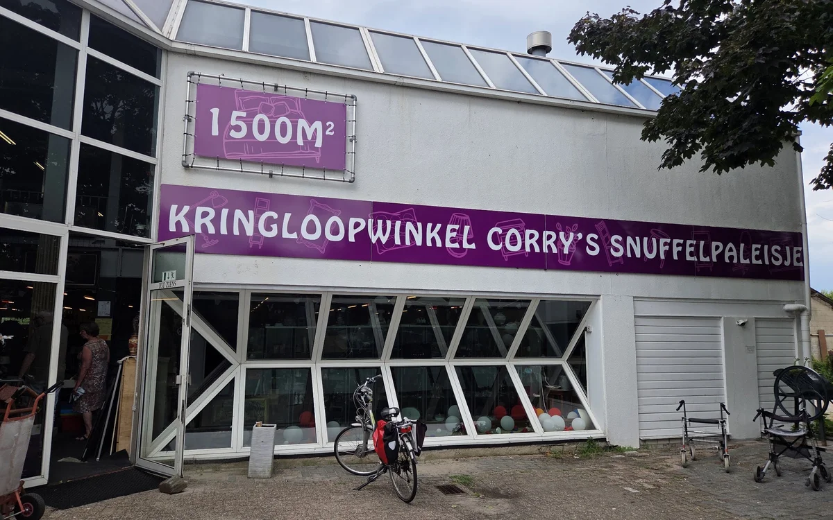 Voorgevel van kringloopwinkel met paars uithangbord en grote raampartijen, fiets ervoor geparkeerd