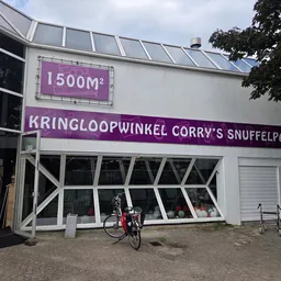 Voorgevel van kringloopwinkel met paars uithangbord en grote raampartijen, fiets ervoor geparkeerd