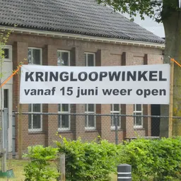 Banner met witte achtergrond en zwarte letters 'KRINGLOOPWINKEL vanaf 15 juni weer open' aan een hek