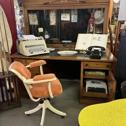 Houten bureau met typemachine, zwarte telefoon en vintage accessoires; oranje-witte bureaustoel ernaast