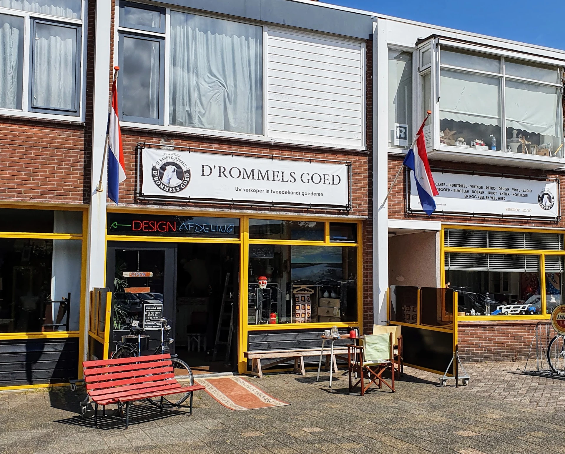 Gevel van kringloopwinkel D'rommels Goed met gele raamkozijnen, rode bank op het trottoir, Nederlandse vlag en winkelnaambord
