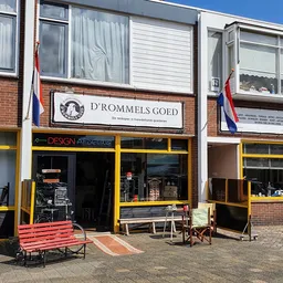Gevel van kringloopwinkel D'rommels Goed met gele raamkozijnen, rode bank op het trottoir, Nederlandse vlag en winkelnaambord