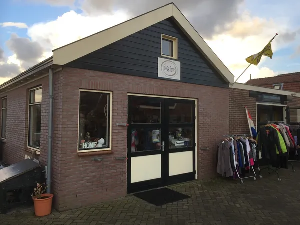 Bakstenen kringloopwinkel met zwarte pui, ramen en ingang; kleding op rekken buiten