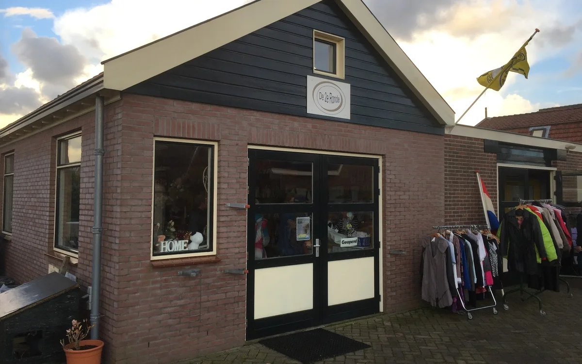 Bakstenen kringloopwinkel met zwarte pui, ramen en ingang; kleding op rekken buiten