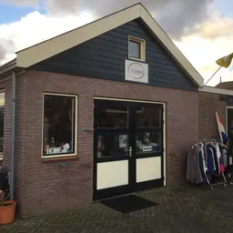 Bakstenen kringloopwinkel met zwarte pui, ramen en ingang; kleding op rekken buiten