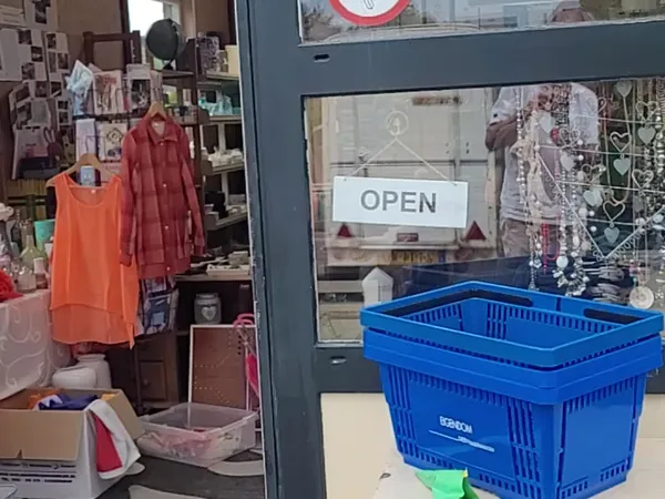Winkelinterieur met kledingrekken, stellingen met spullen, blauw winkelmandje op toonbank en OPEN-bord in raam