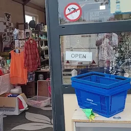 Winkelinterieur met kledingrekken, stellingen met spullen, blauw winkelmandje op toonbank en OPEN-bord in raam