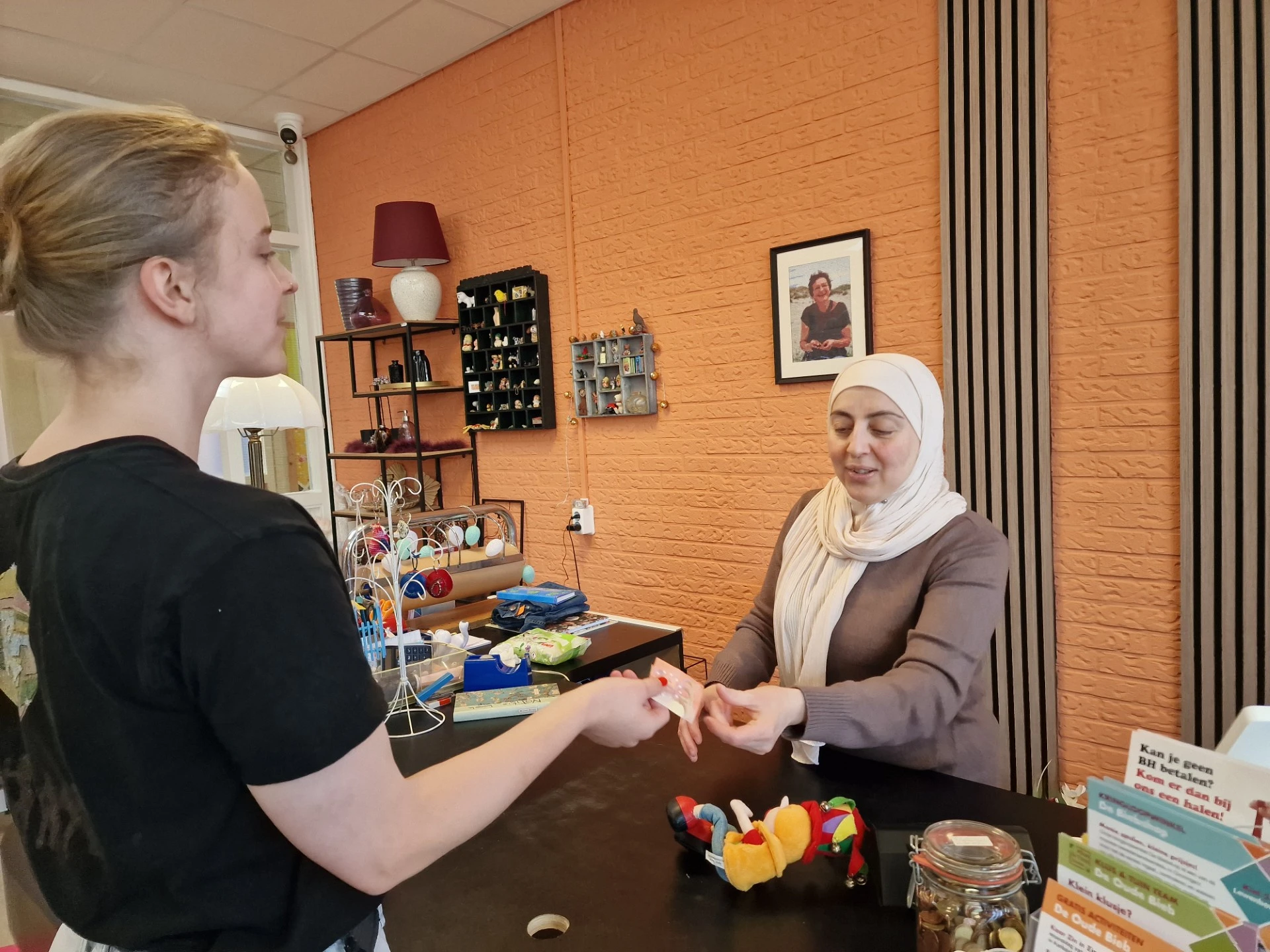 Twee vrouwen achter een toonbank in een winkel met oranje muur, één helpt de ander met iets in haar handen