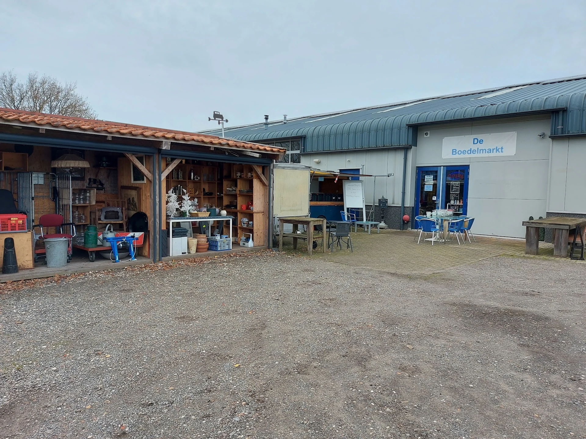Voorkant kringloopwinkel met houten afdak links, blauwe luifel rechts en 'De Boedelmarkt' zichtbaar op de witte gevel