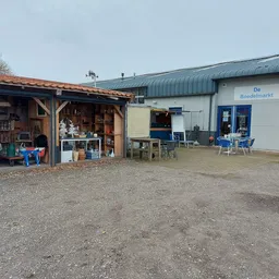 Voorkant kringloopwinkel met houten afdak links, blauwe luifel rechts en 'De Boedelmarkt' zichtbaar op de witte gevel