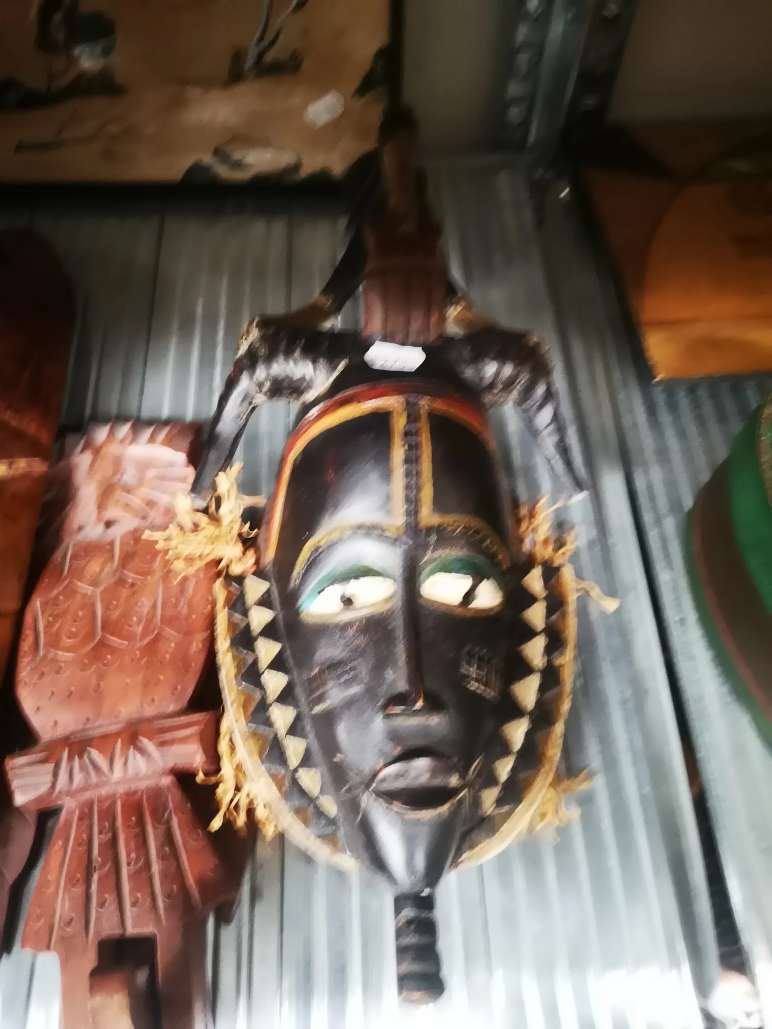 Zwart Afrikaans houten masker met goudkleurige decoratie en veren, aan een grijze stof opgehangen