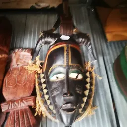 Zwart Afrikaans houten masker met goudkleurige decoratie en veren, aan een grijze stof opgehangen