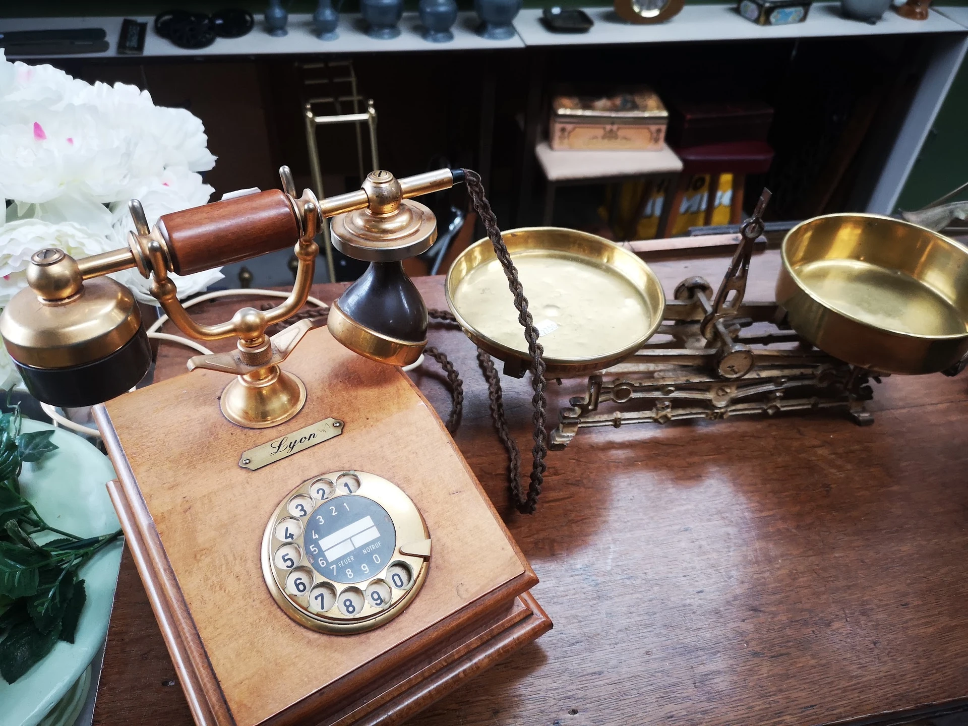 Vintage telefoon met houten basis en gouden bel, naast koperen weegschaal en andere antieke huishoudartikelen op houten tafel