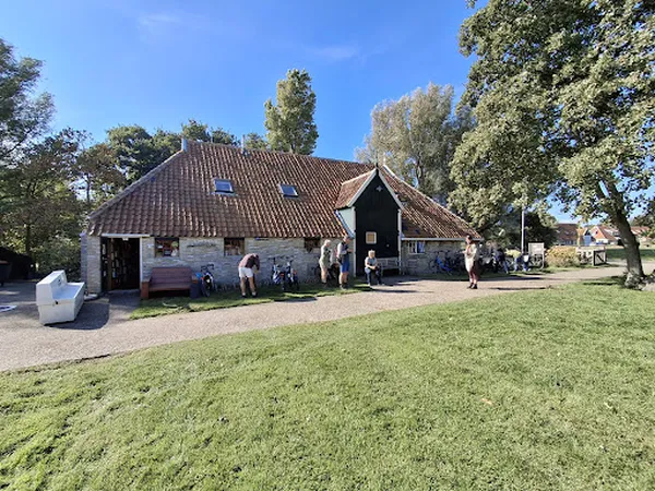 Voorkant van een wit kringloopwinkelgebouw met donker dak en enkele bezoekers buiten, omgeven door groen.