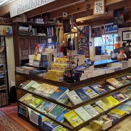 Boekenwinkel interieur met toonbank, boeken op stellingen en in vitrines, gele dozen en diverse artikelen.