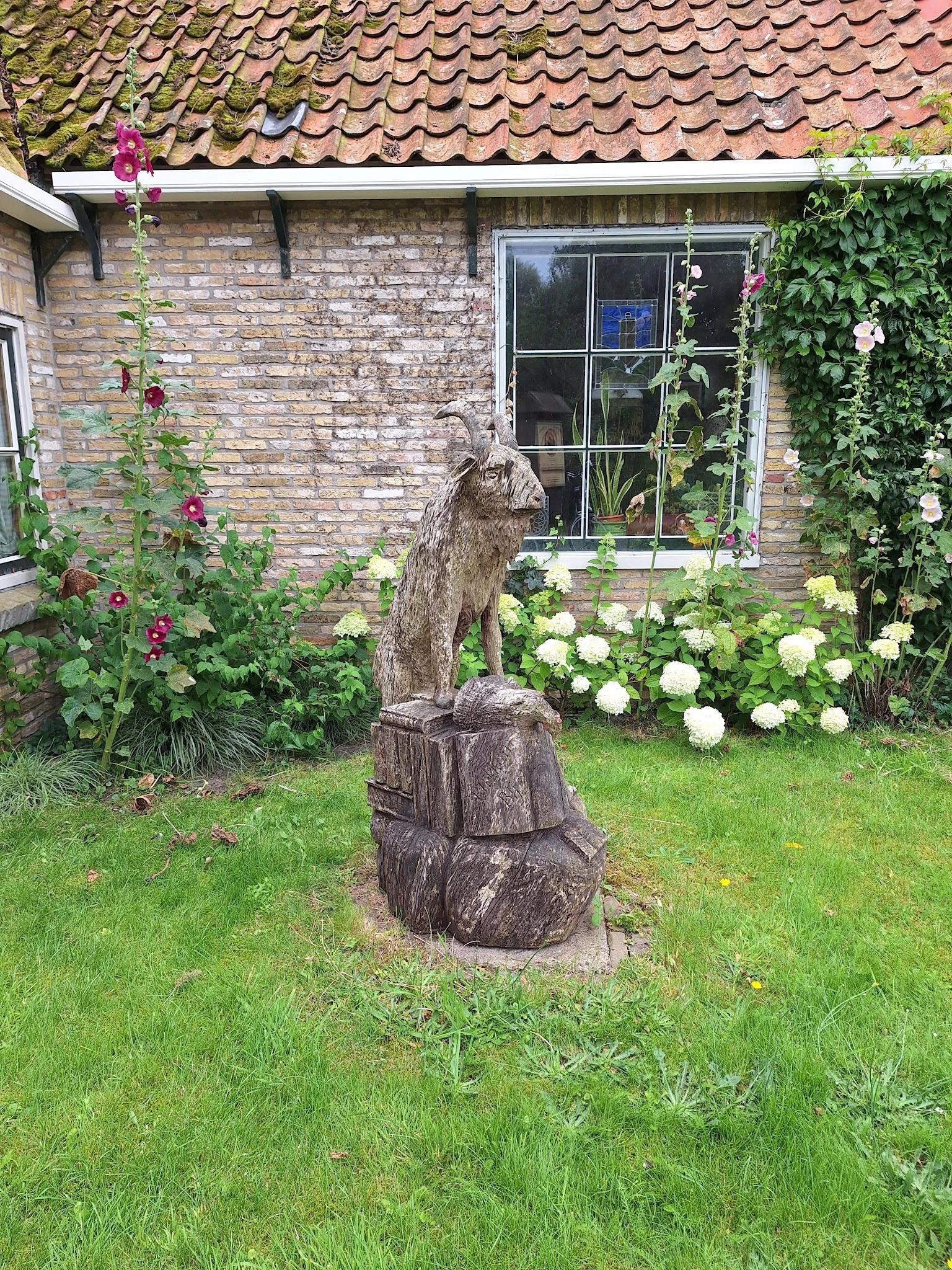 Houten tuinbeeldje van een kat op sokkel tussen bloeiende hortensia's tegen bakstenen huis