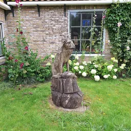 Houten tuinbeeldje van een kat op sokkel tussen bloeiende hortensia's tegen bakstenen huis