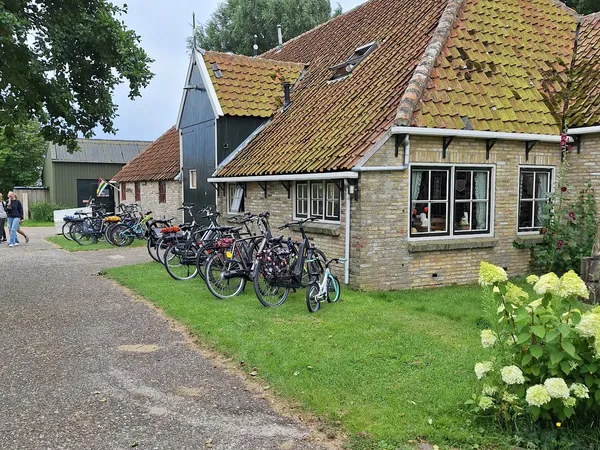 Voorkant van stenen kringloopwinkel met gele dakpannen, fietsen geparkeerd op het gazon, groene hortensia's op de voorgrond