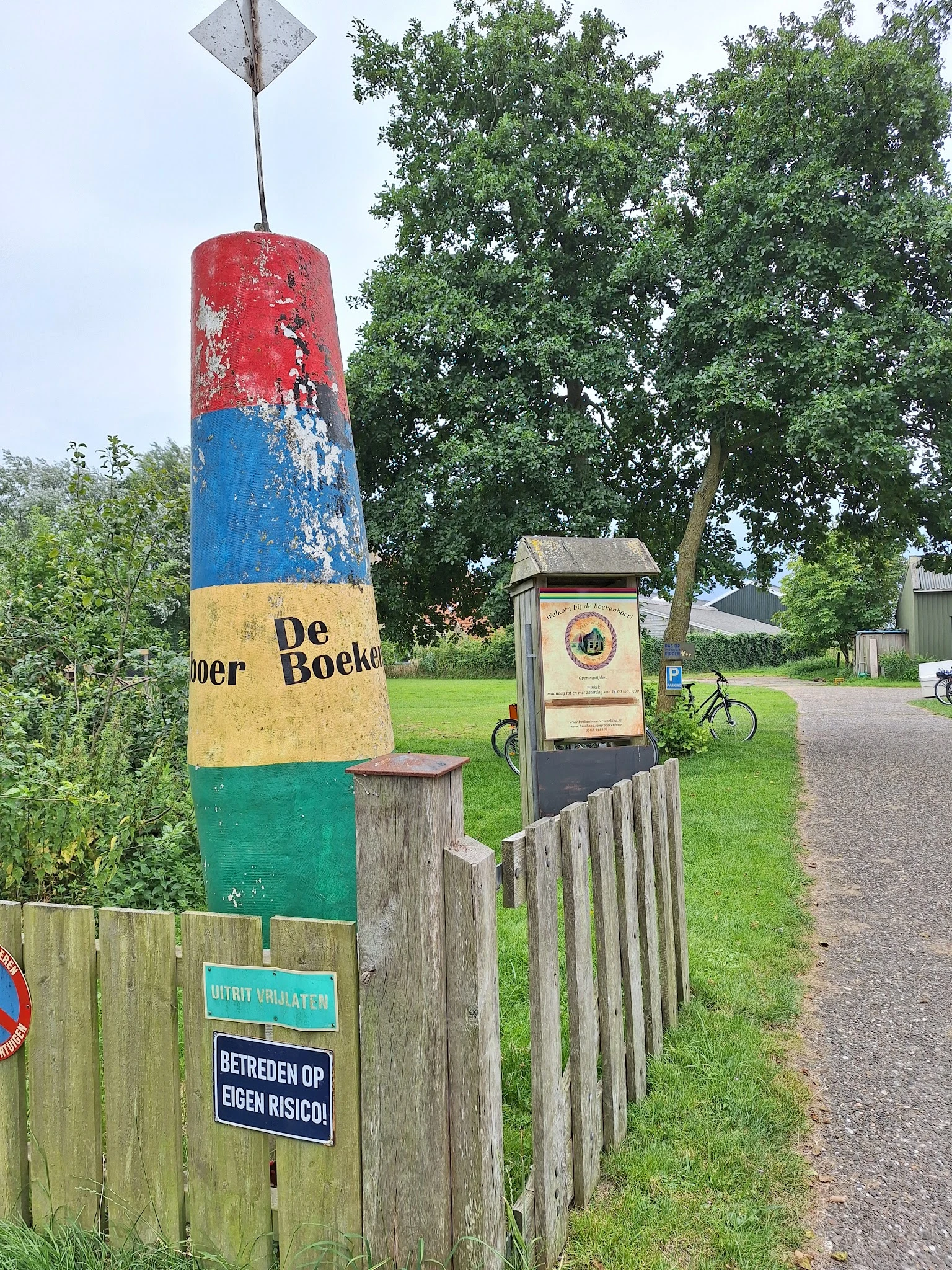 Gekleurde paal met 'De Boekenboer' tekst en winkelingang met houten hek langs landelijk pad