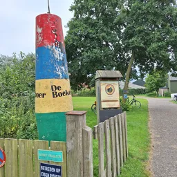 Gekleurde paal met 'De Boekenboer' tekst en winkelingang met houten hek langs landelijk pad