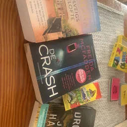 Boekenrek met tweedehands boeken, waaronder 'De Crash' en andere titels, en enkele speelgoed- of puzzeldozen