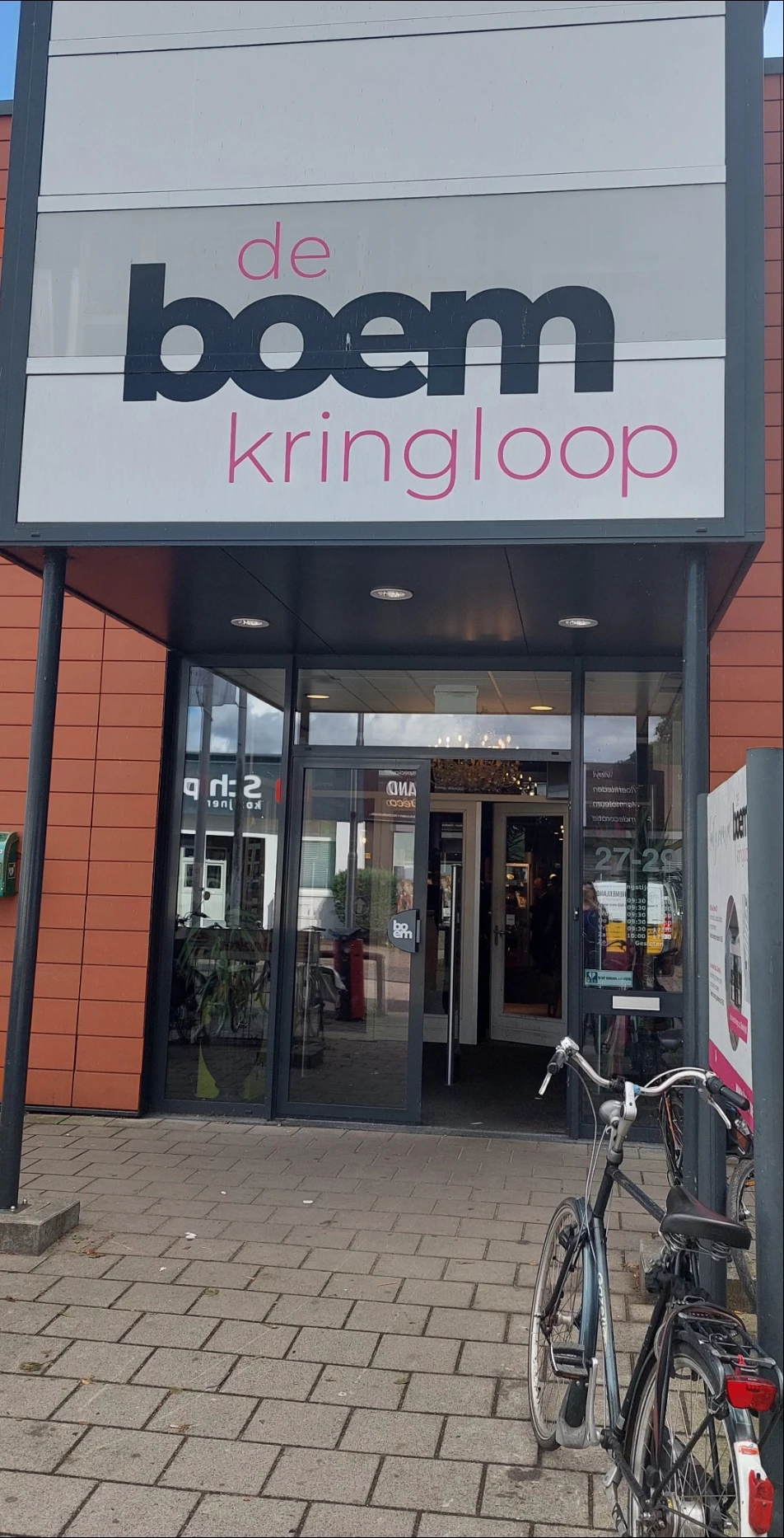 Glazen pui van kringloopwinkel met zwart frame en ingang, rode bakstenen gevel