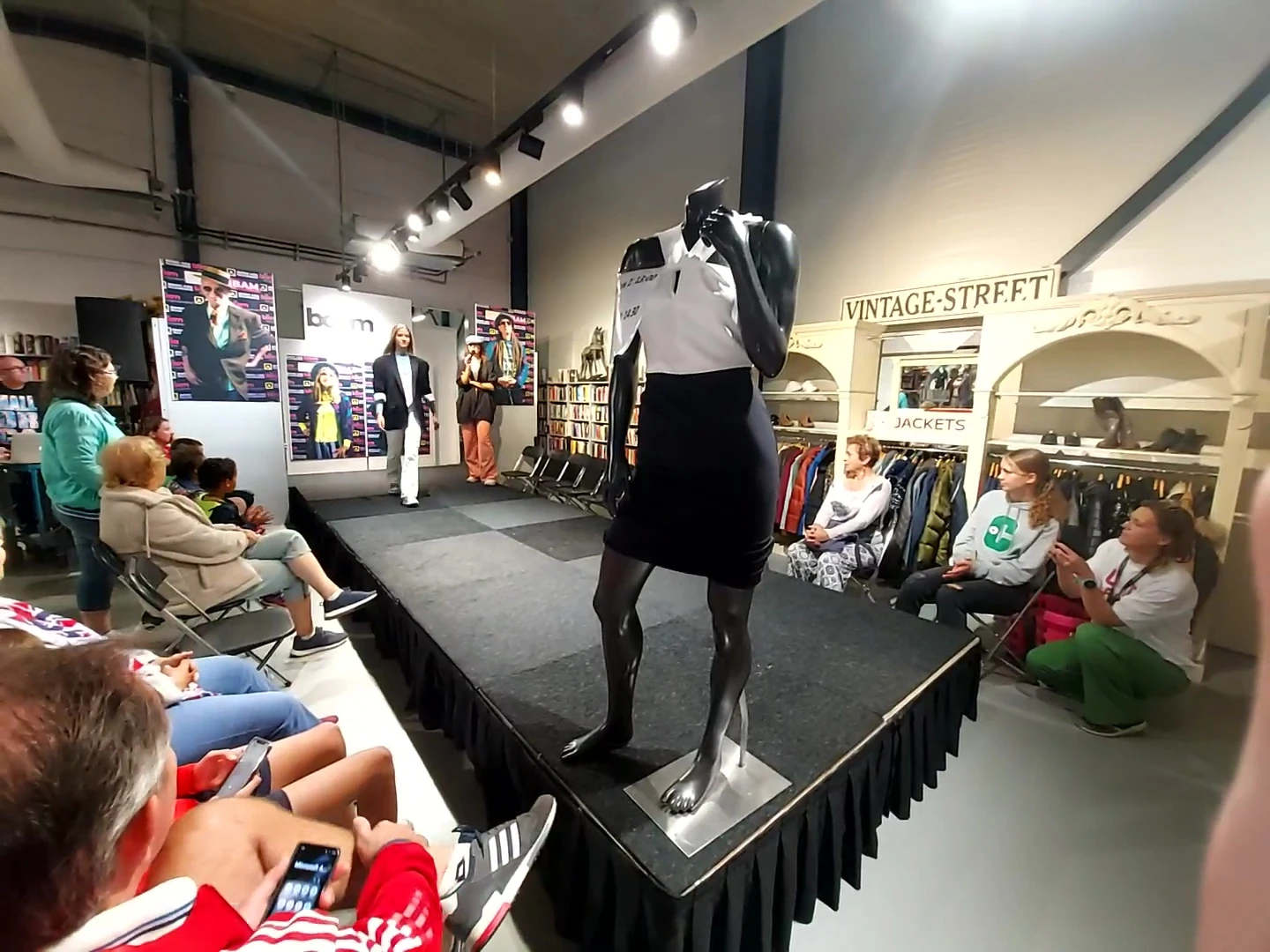 Binnenaanzicht van kringloopwinkel met modepop op podium, bezoekers kijken naar modeshow, kledingrekken en vintage-spullen zi