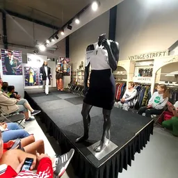 Binnenaanzicht van kringloopwinkel met modepop op podium, bezoekers kijken naar modeshow, kledingrekken en vintage-spullen zi