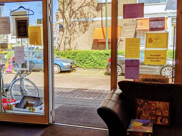 Winkelinterieur met uitzicht door glazen deur naar straat, fiets zichtbaar links, gele en rode borden in raam