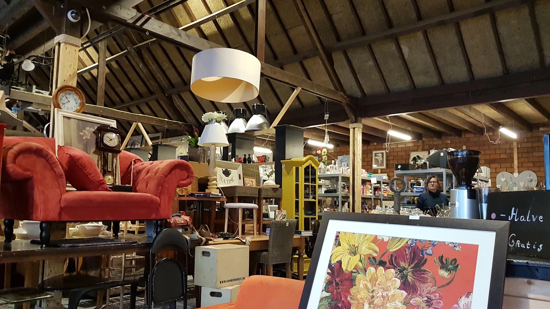Ruim interieur van kringloopwinkel met houten spanten, rode fauteuil, gele kast, diverse meubels en artikelen, moderne hangla