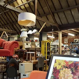 Ruim interieur van kringloopwinkel met houten spanten, rode fauteuil, gele kast, diverse meubels en artikelen, moderne hangla