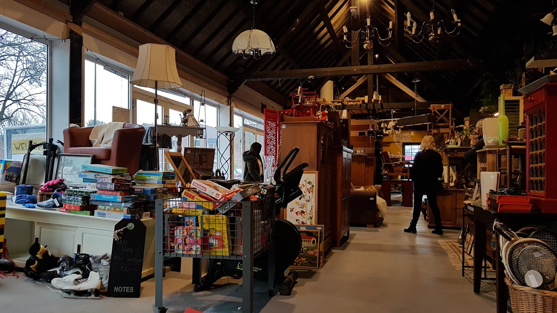 Ruim interieur van kringloopwinkel met diverse meubelen, boeken, decoratiespullen en klanten die aan het winkelen zijn