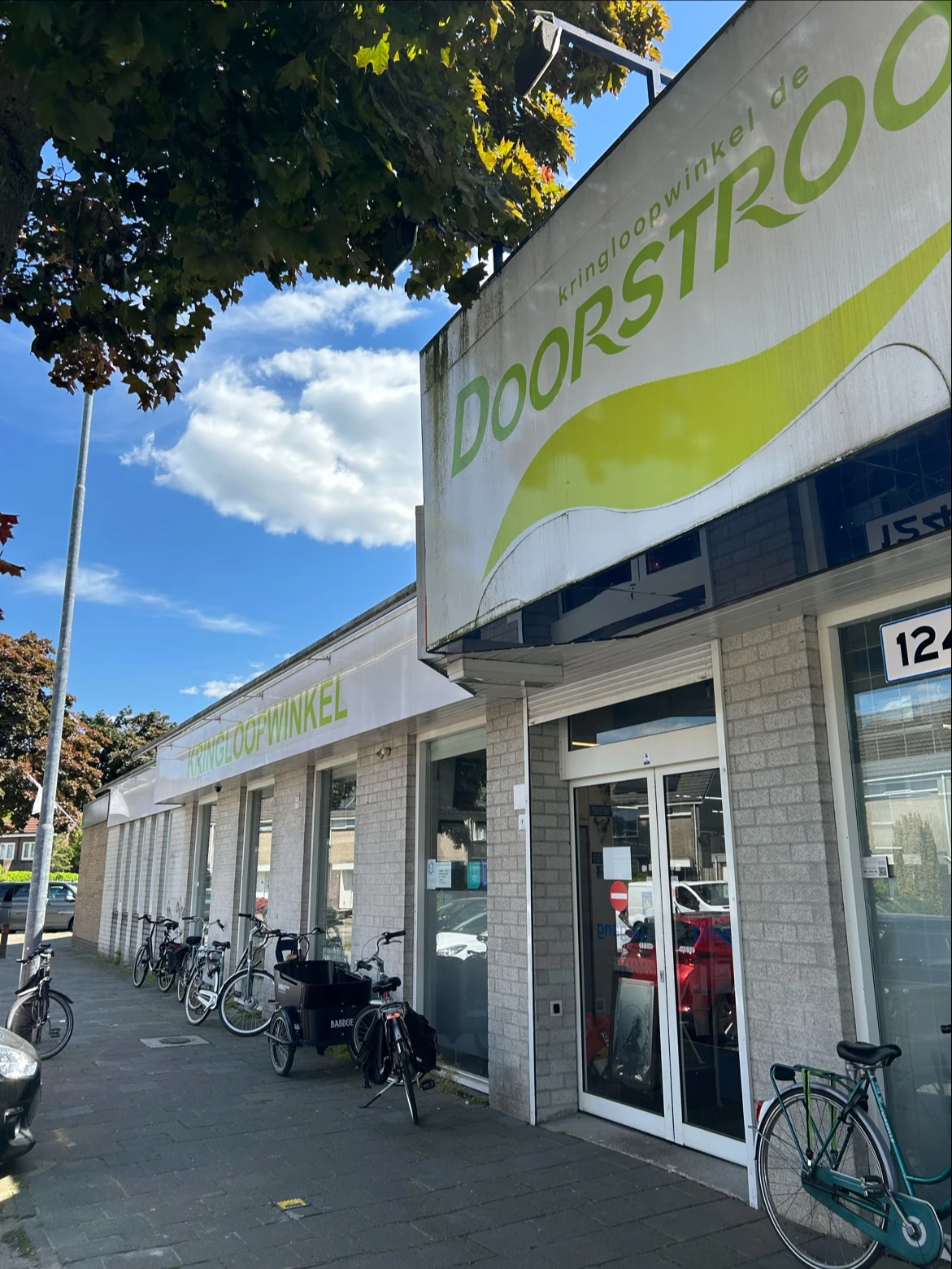 Grijze bakstenen gevel van kringloopwinkel met groene logo en etalageruiten, fietsen geparkeerd ernaast