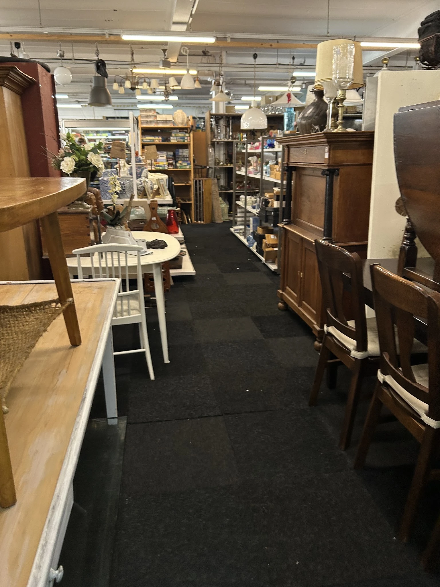 Winkelinterieur met houten meubels, witte stoelen en zwarte looppad tussen stellingen met diverse spullen