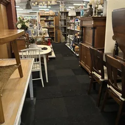 Winkelinterieur met houten meubels, witte stoelen en zwarte looppad tussen stellingen met diverse spullen